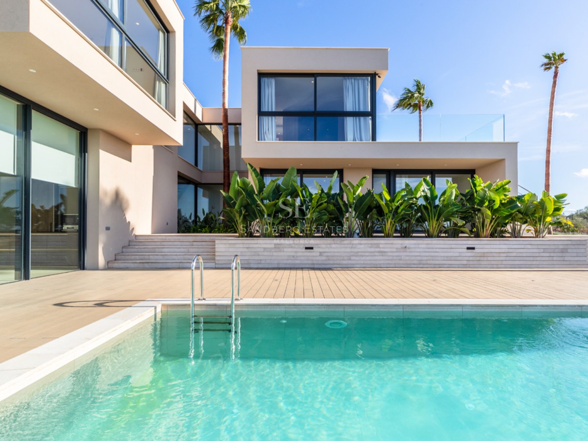 Modern beige villa med stora fönsterpartier mot en kristallklar pool och palmer under blå himmel.