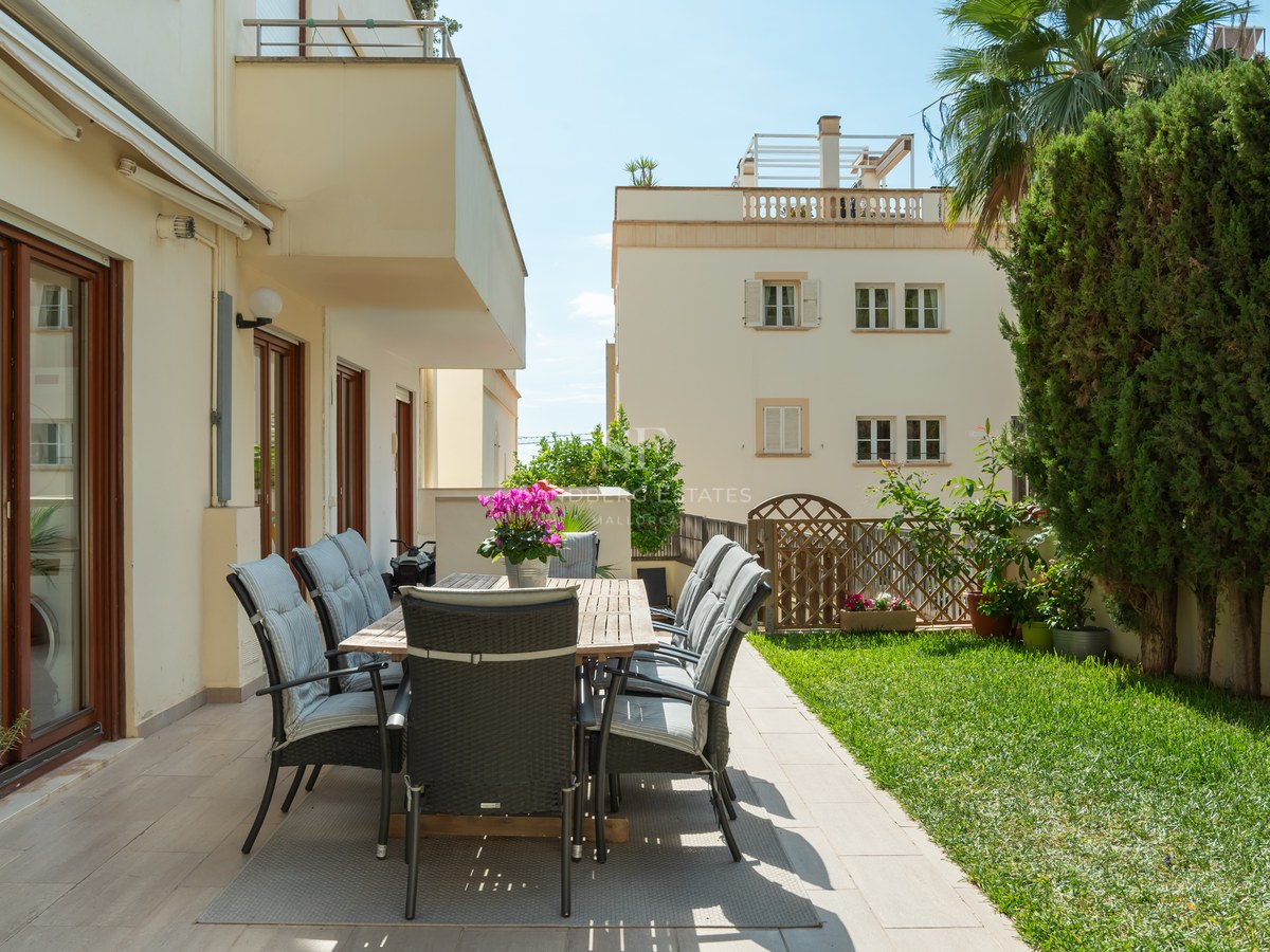 Comedor exterior en terraza de baldosas junto a jardín con césped y setos altos bajo luz solar directa.