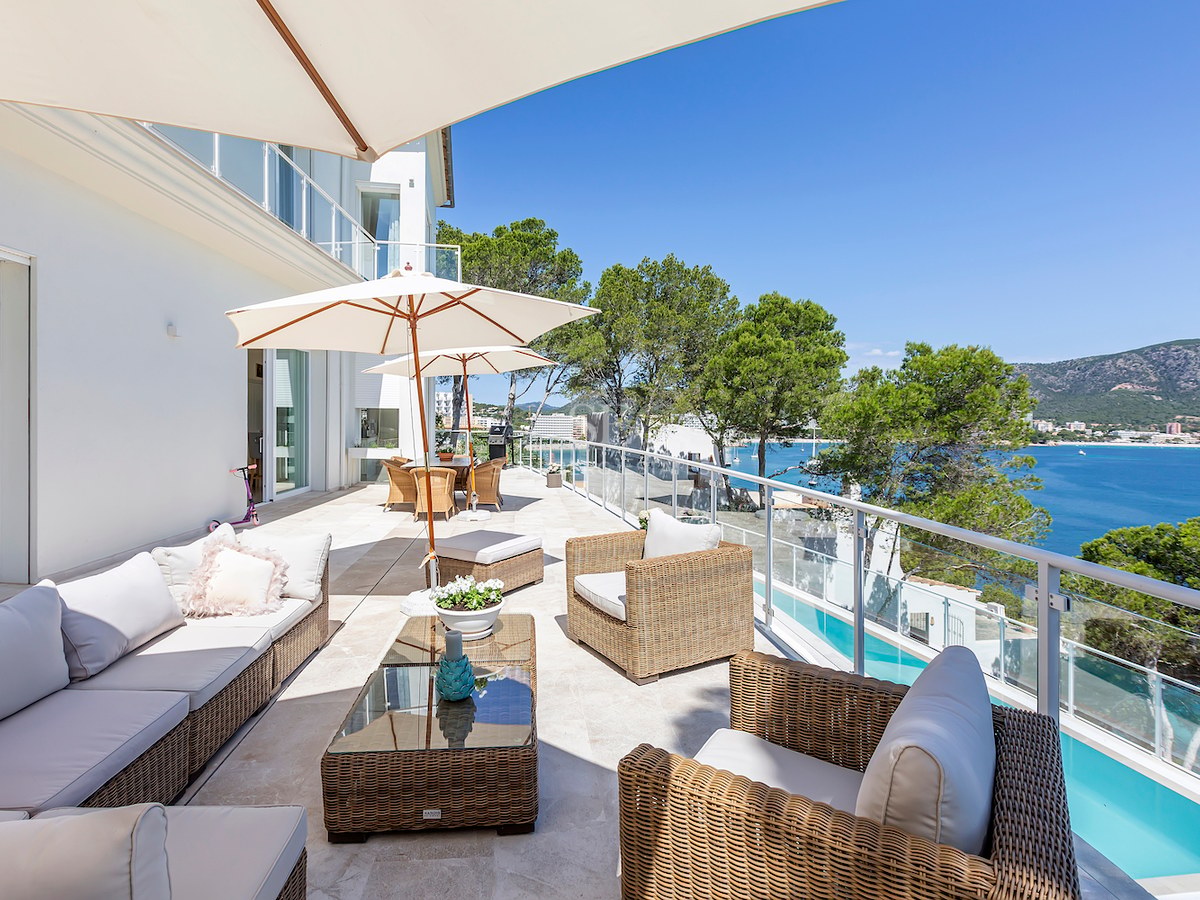 Spacious white stone terrace with wicker furniture, glass railings, and a panoramic view of the sea and mountains.