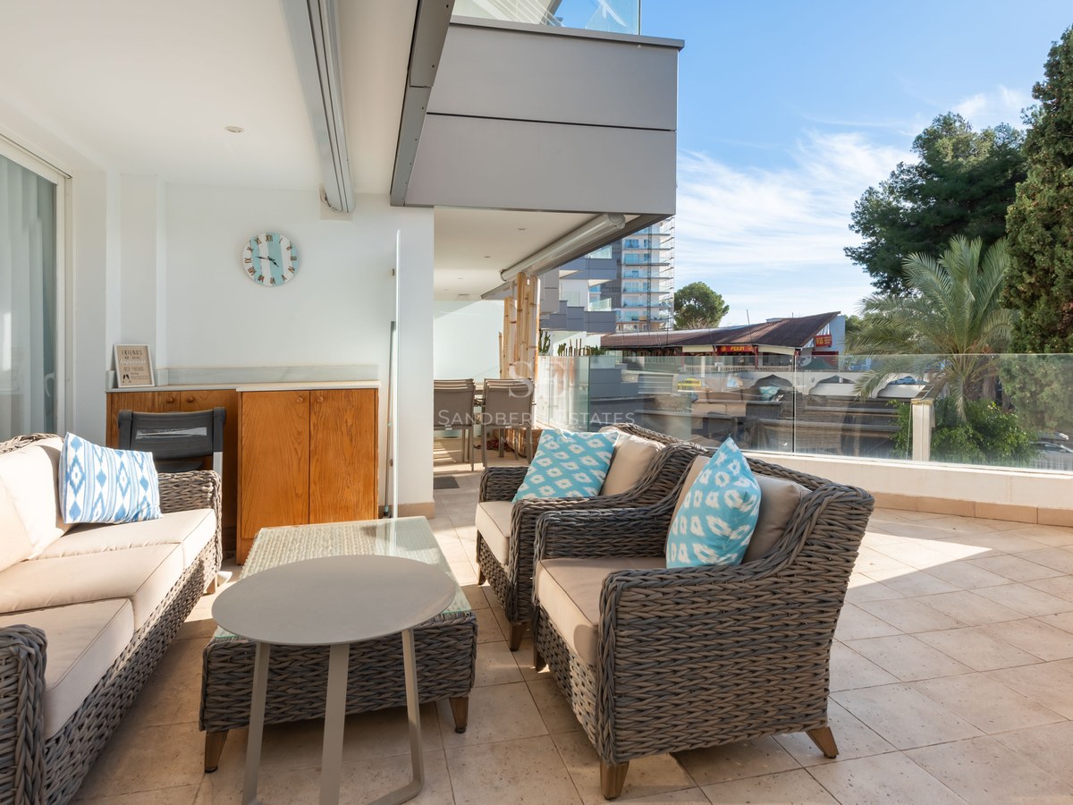 Spacious terrace with grey wicker sofas, blue accent cushions, and glass railings under a clear blue sky.