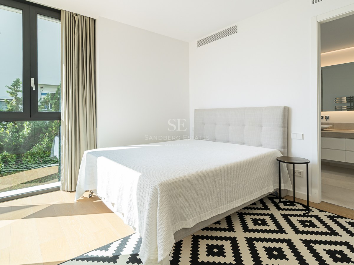 Chambre moderne avec lit blanc, tapis géométrique, grande fenêtre sur jardin et salle de bain attenante.