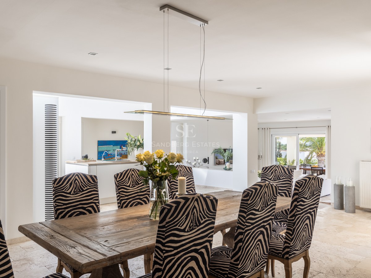 Table à manger en bois rustique, chaises à motif zébré et sols en travertin dans une villa lumineuse.