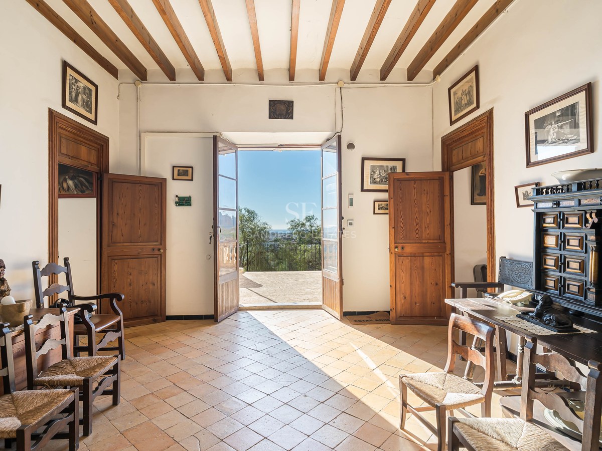 Vestíbulo tradicional con vigas de madera, suelo de terracota y puertas abiertas hacia una terraza soleada.