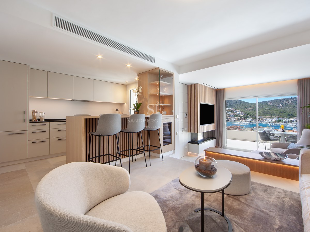 A bright, modern open-plan living room and kitchen featuring beige cabinetry, an island with stools, and a large window with a marina view.
