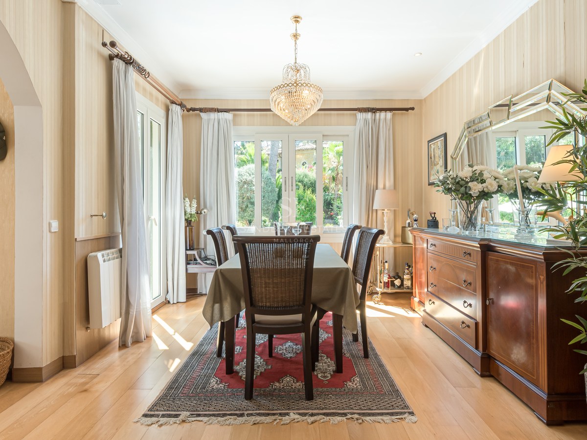Salle à manger ensoleillée avec table en bois, lustre en cristal et grand buffet donnant sur l'extérieur.