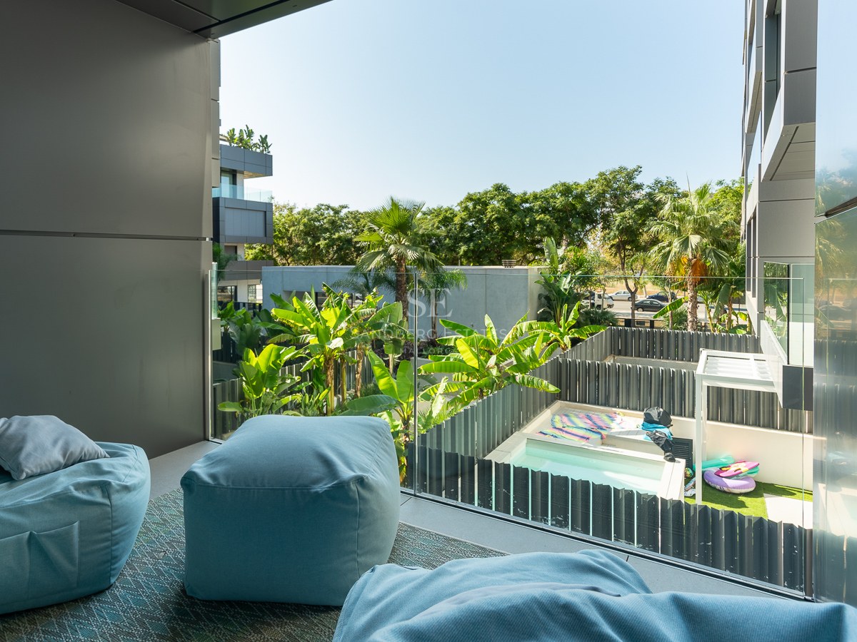 Balcon moderne avec poufs bleu clair et garde-corps en verre surplombant un jardin tropical et une piscine.