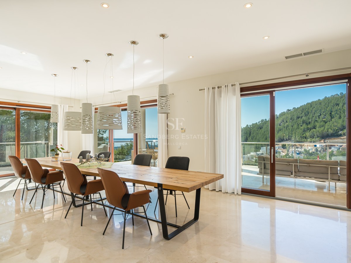 Comedor moderno con mesa de madera, suelo de mármol y ventanales con vistas a la montaña y el mar.
