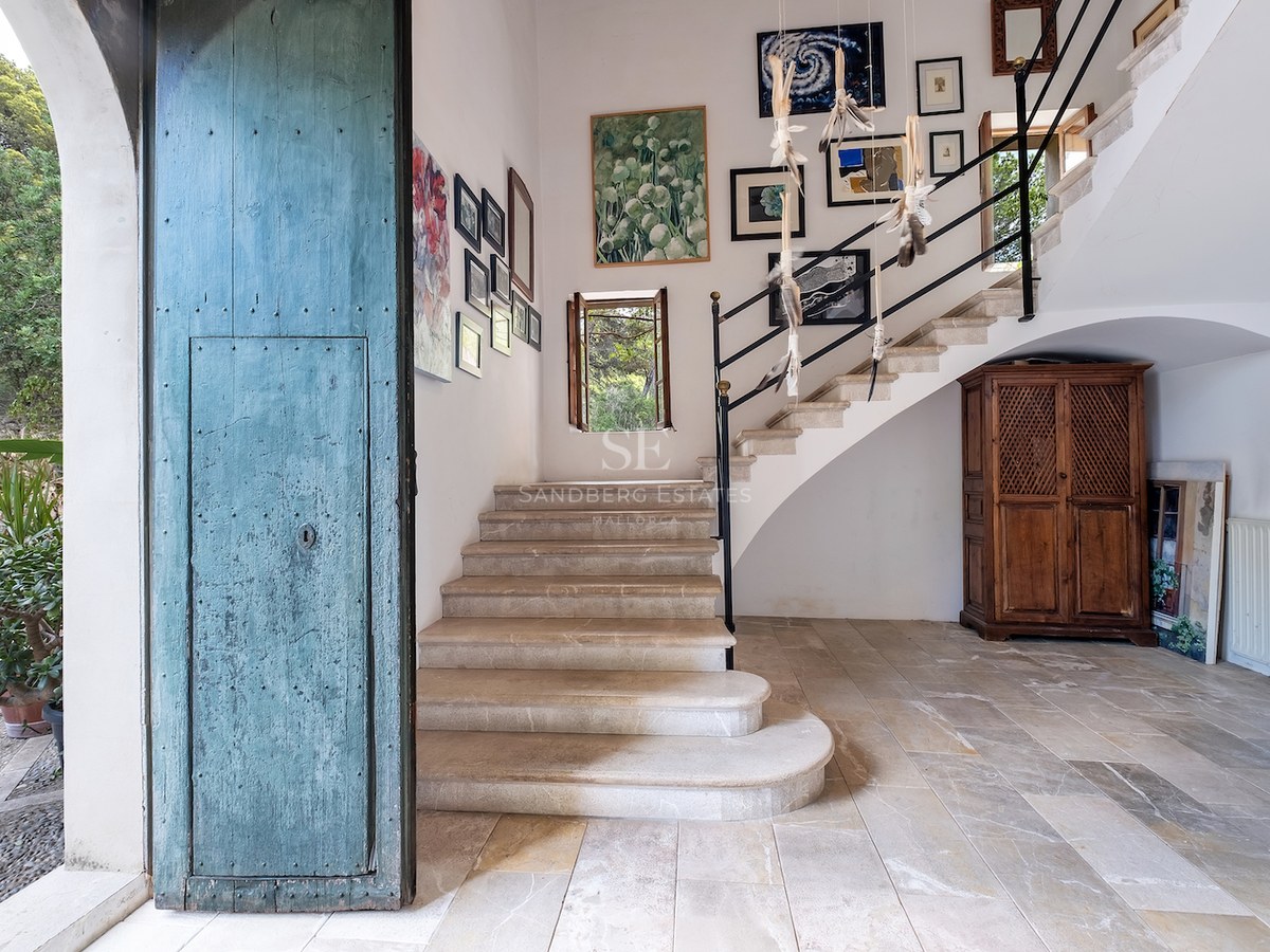 Hall d'entrée avec une grande porte bleue vieillie, escalier en pierre et galerie de tableaux sur murs blancs.