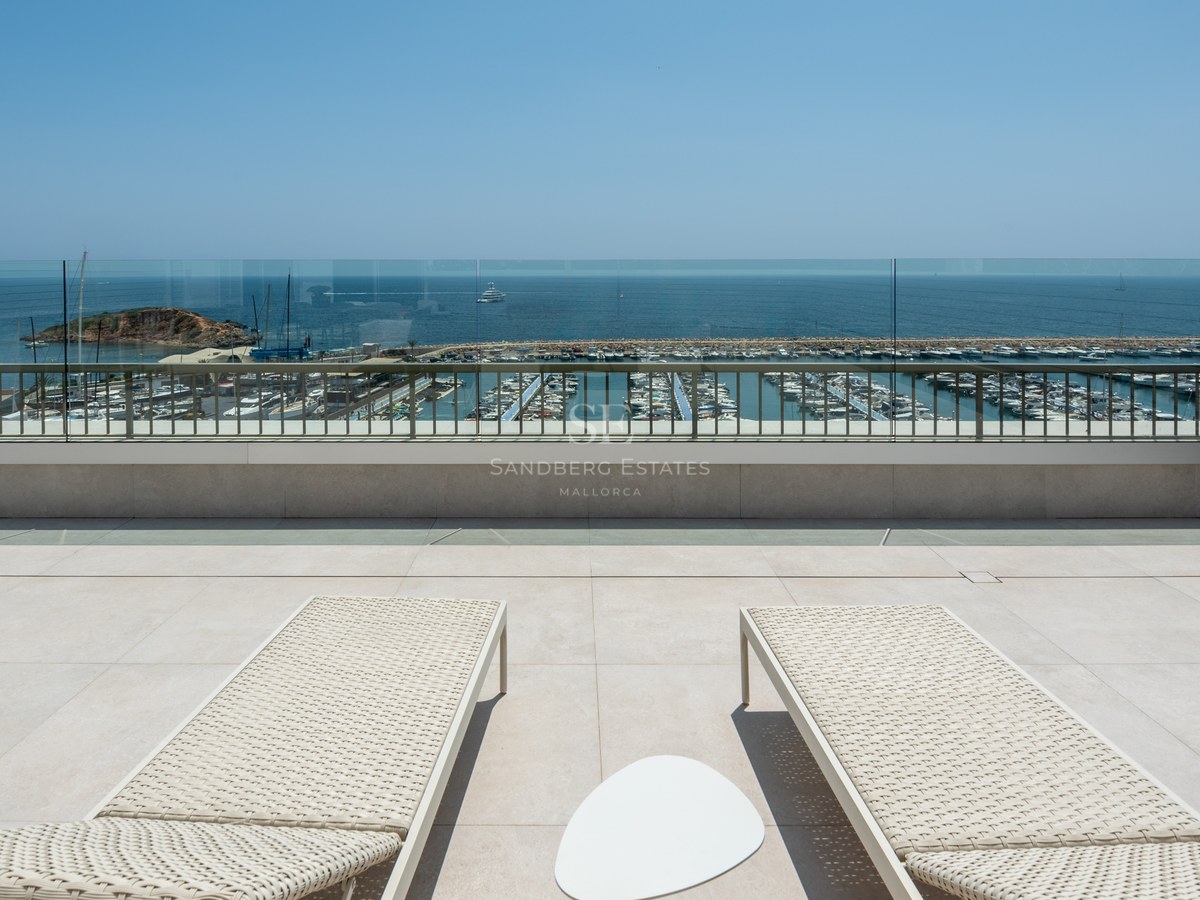 Two cream sun loungers on a stone terrace overlooking a yacht marina and the Mediterranean Sea under a clear sky.