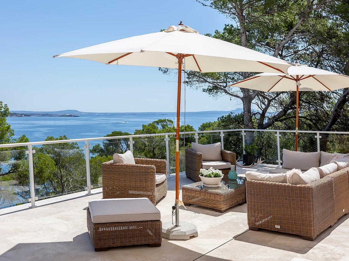 Sun-drenched terrace with wicker lounge furniture, white umbrellas, and glass railings overlooking the blue sea.