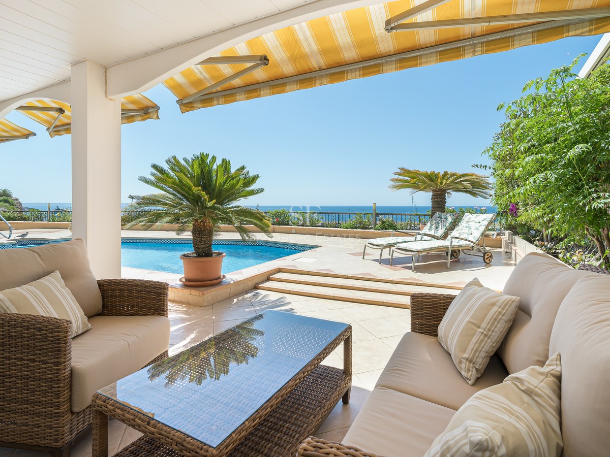 Covered terrace with wicker sofas facing a blue pool and the sea under a clear blue sky.