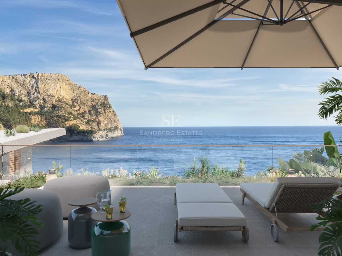 A sunny terrace with sun loungers, a parasol, and a glass railing overlooking the Mediterranean sea and a rocky cliff.