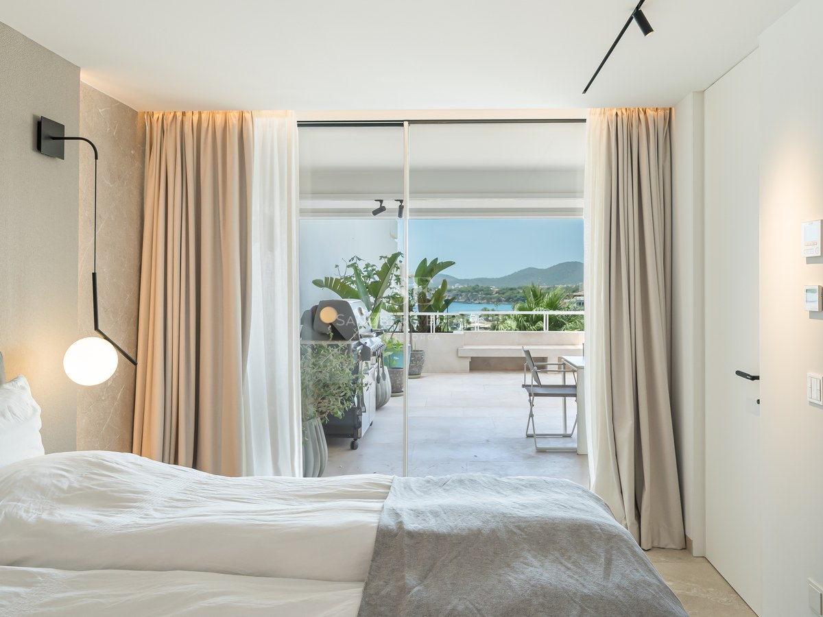 Chambre moderne avec lit blanc et portes vitrées s'ouvrant sur une terrasse avec barbecue et vue sur la mer.