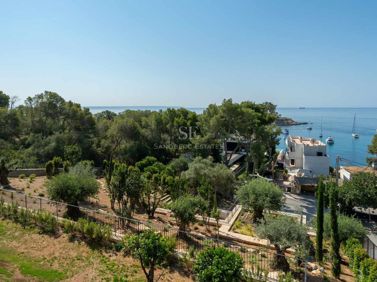 Elevated view of a Mediterranean garden with olive trees overlooking a blue bay with sailboats and white houses.