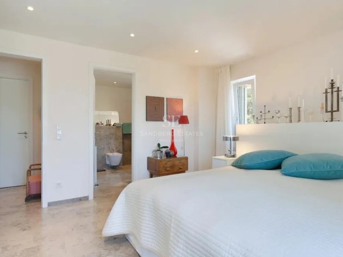 Modern bedroom featuring white bedding, turquoise pillows, marble floors, and an open doorway to a stone bathroom.