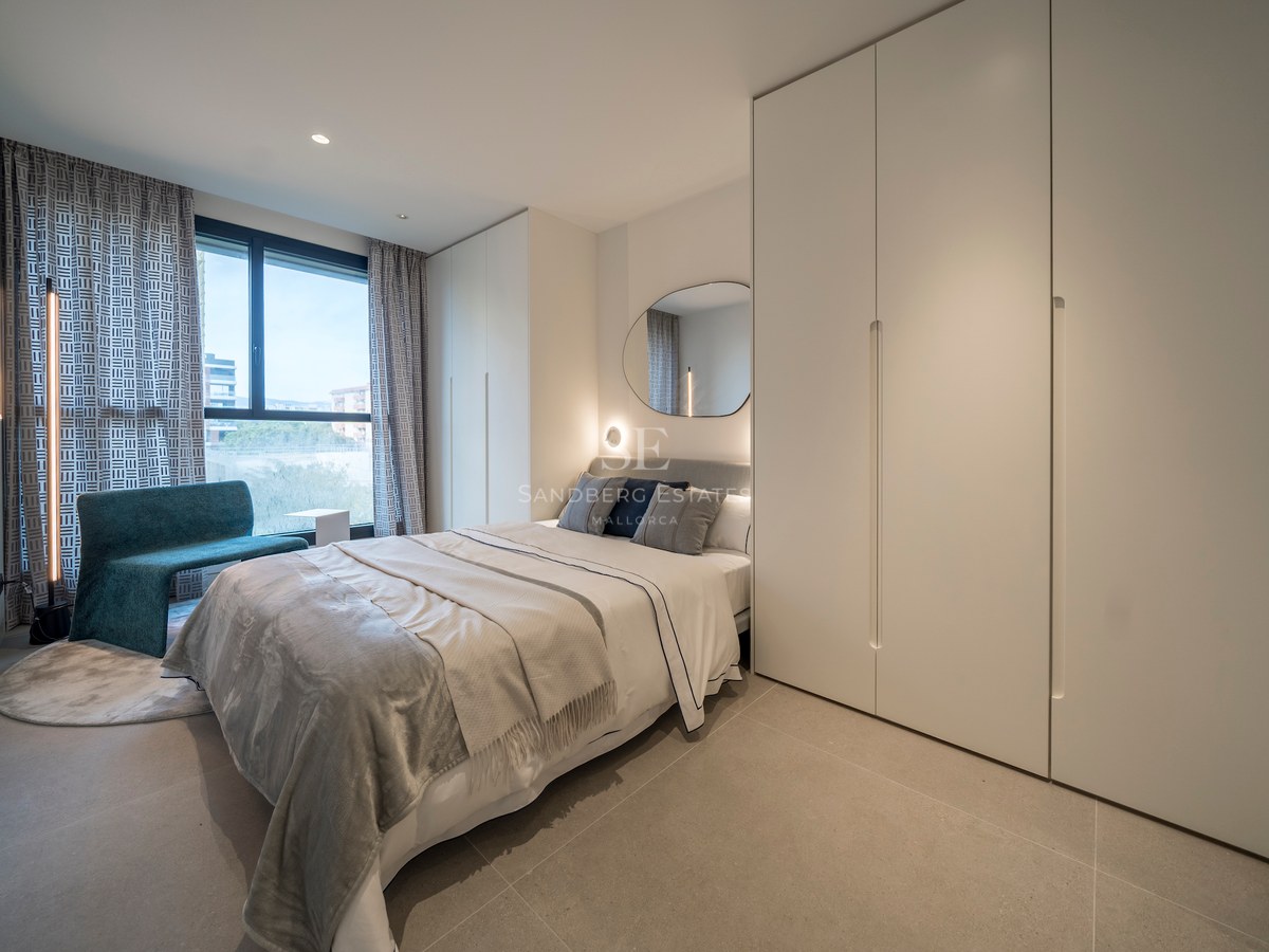 Une chambre moderne lumineuse avec un lit gris, de grandes armoires blanches, une chaise sarcelle et une grande fenêtre.