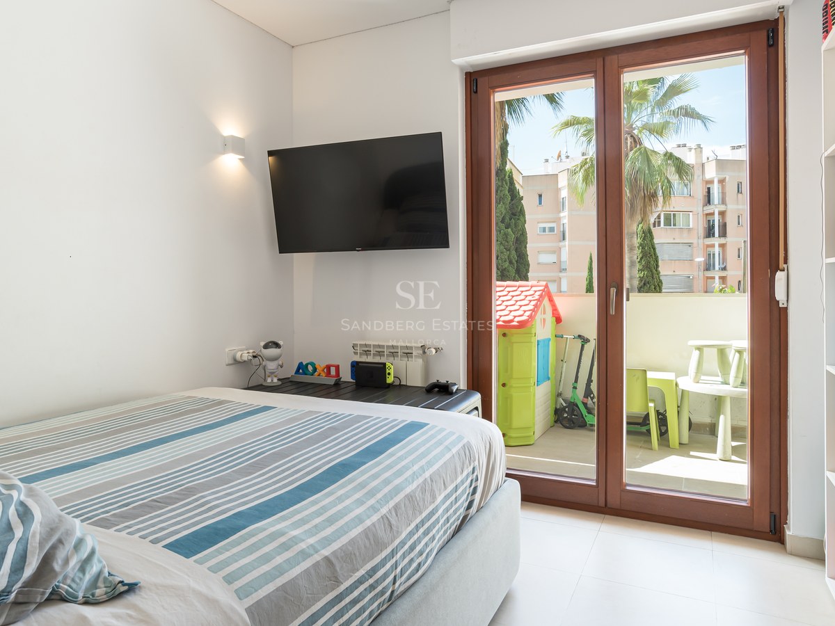 Dormitorio luminoso con cama de rayas, TV en pared y puertas de cristal que dan a una terraza con juegos infantiles.