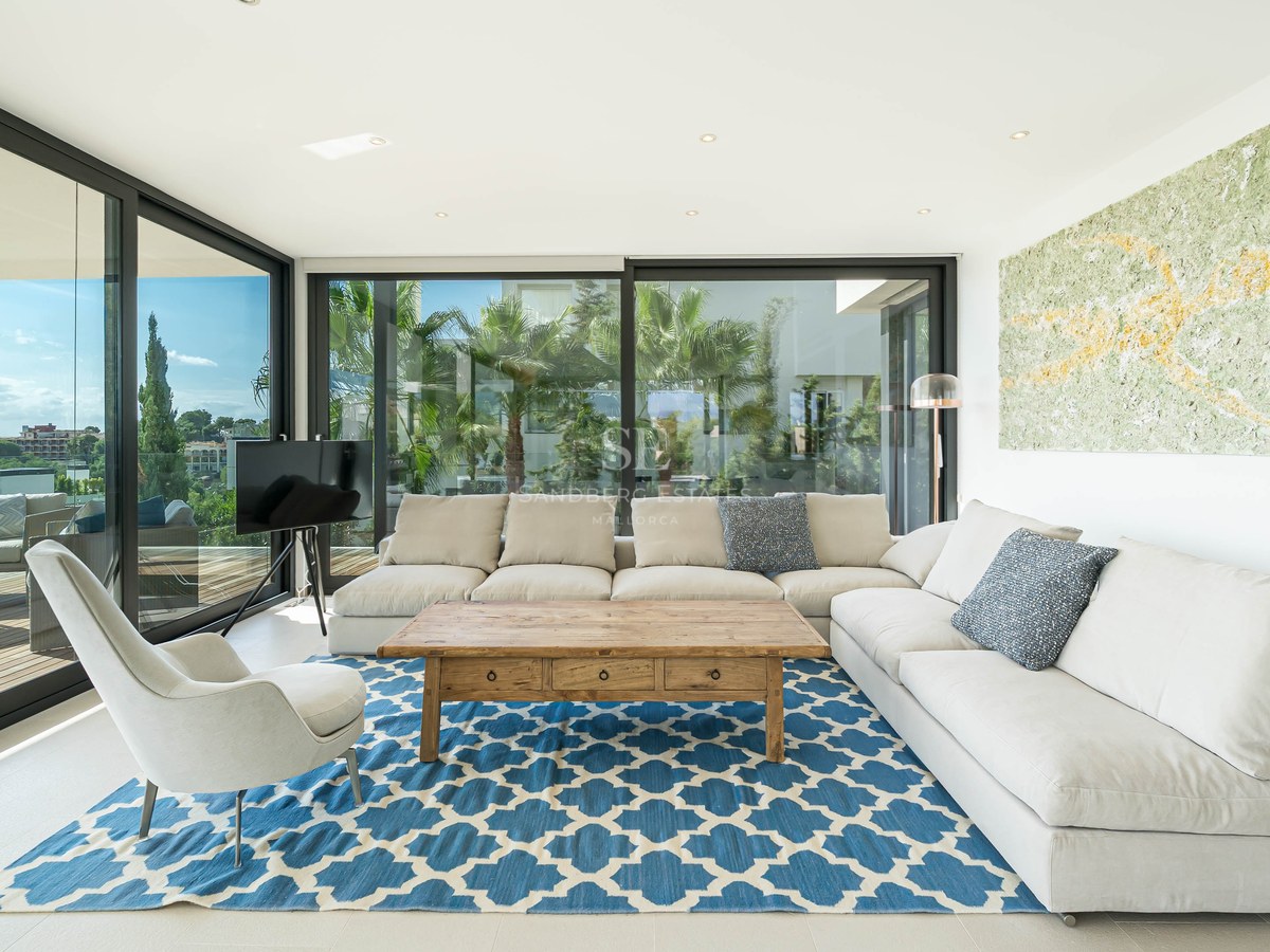 Salon moderne avec canapé en L blanc, tapis bleu à motifs et baies vitrées offrant une vue sur la côte.