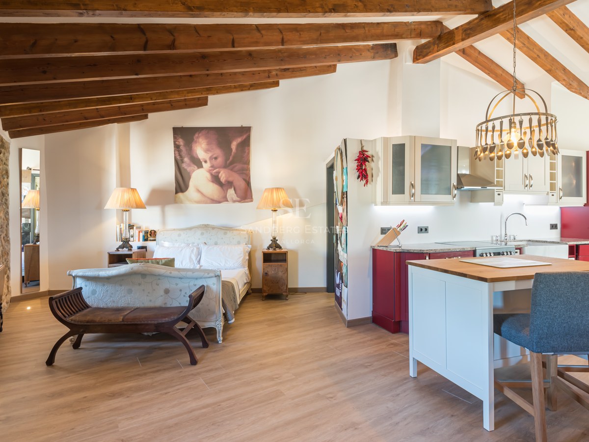 Studio avec lit classique, poutres en bois apparentes, mur en pierre et cuisine rouge et blanche avec îlot.