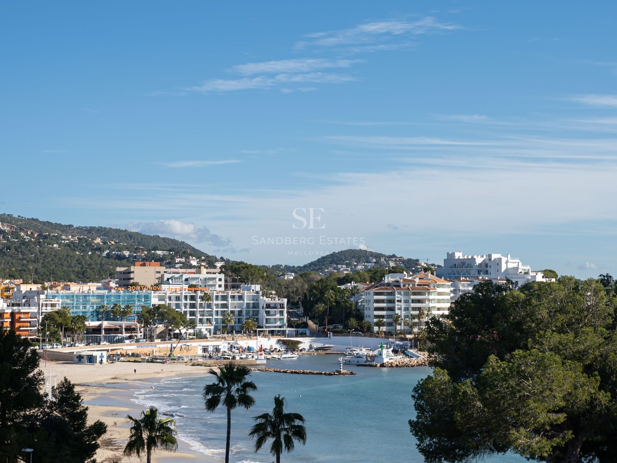 A wide view of a coastal town with white buildings, a sandy beach, palm trees, and a small harbor under a clear blue sky.