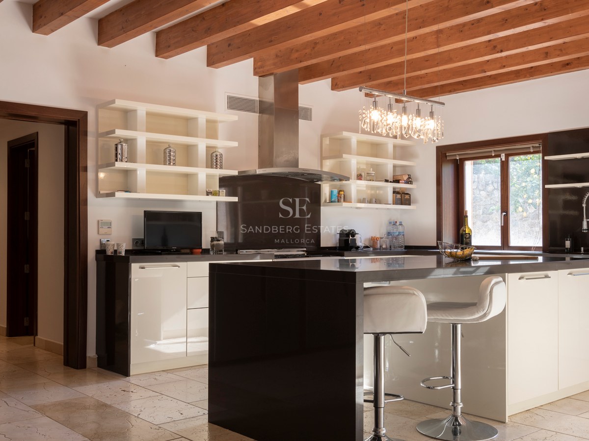 Cuisine moderne avec îlot noir, placards blancs, poutres en bois apparentes et sol en pierre naturelle.