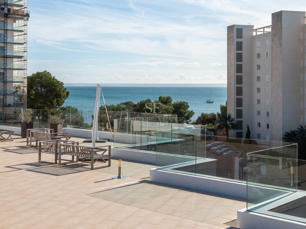 Spacious tiled terrace with glass railings and wooden seating, overlooking the blue sea and coastal buildings.