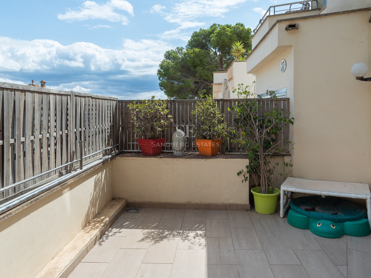 Terraza privada con suelo cerámico, valla de madera, plantas en macetas y vistas a los árboles bajo un cielo azul.