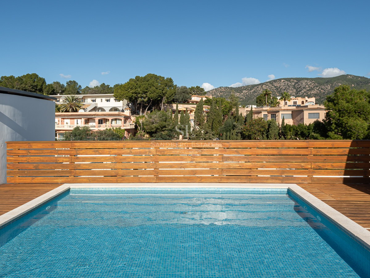 A rectangular rooftop swimming pool with blue tiles and a wooden deck, overlooking green hills and houses.