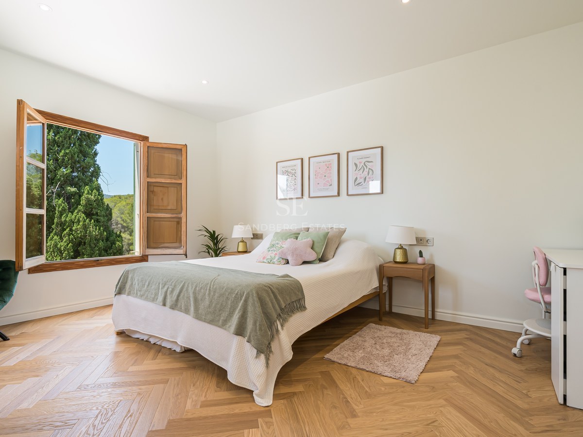Bright bedroom featuring oak herringbone floors, a green velvet chair, and an open window looking onto lush trees.