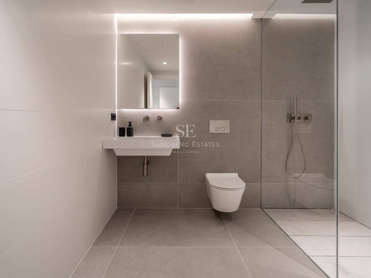 Salle de bain moderne avec carrelage gris, miroir rétroéclairé, lavabo blanc, WC suspendu et paroi de douche en verre.