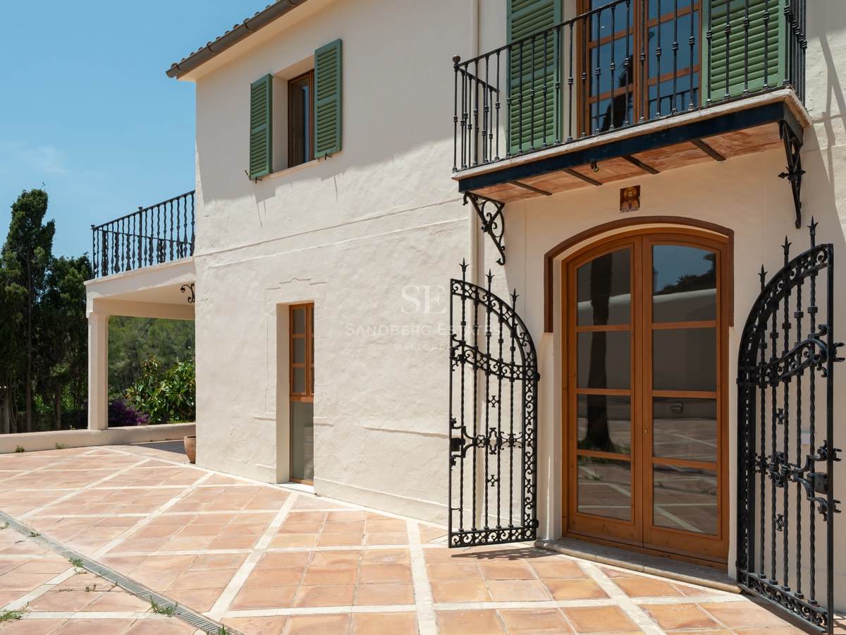 Cream stucco villa with green shutters, wrought iron balcony, and terracotta terrace under a clear blue sky.