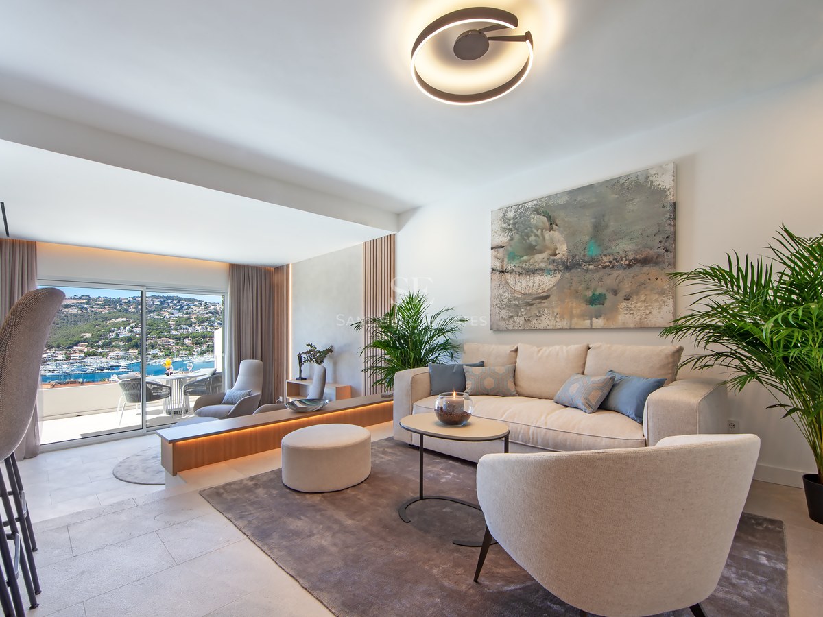 Bright modern living room with cream furniture, large windows overlooking the sea, and lush indoor plants.