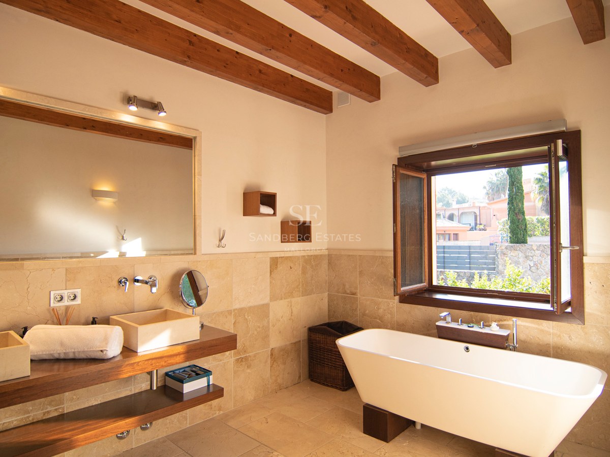 Salle de bains avec poutres apparentes, baignoire îlot, double vasque en pierre et vue sur le jardin.