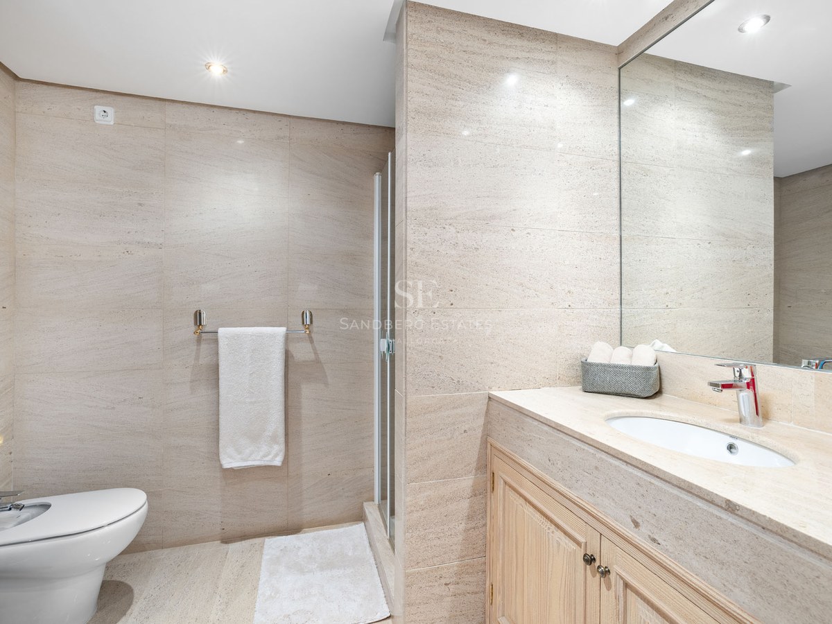 Salle de bain moderne avec murs en pierre beige, bidet blanc, meuble en bois avec lavabo et grand miroir.