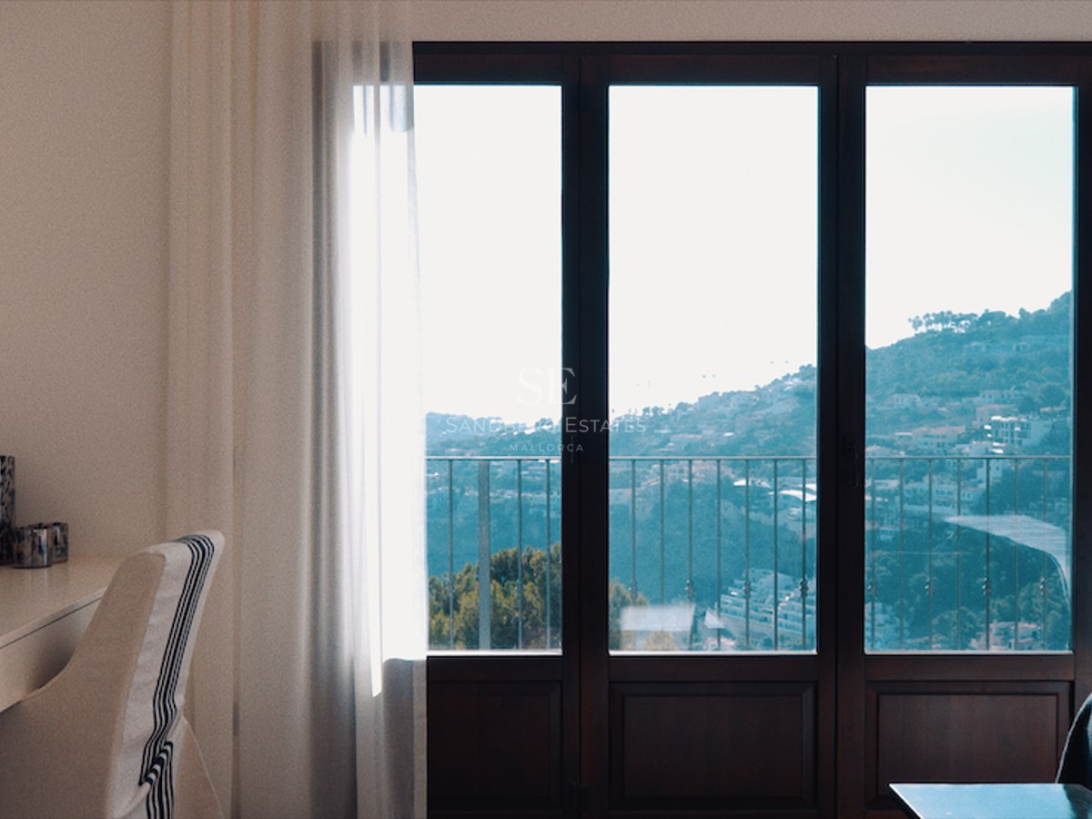 A modern bedroom featuring a desk, a wall-mounted TV, and floor-to-ceiling windows overlooking a scenic hillside and sea.