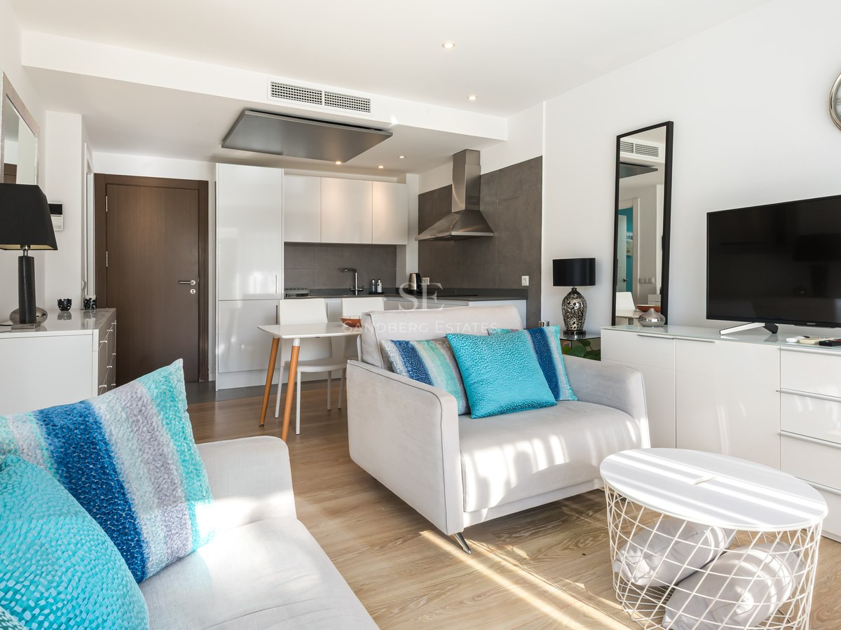 Bright living room with grey armchair, teal cushions, TV console, and modern kitchen in the background.