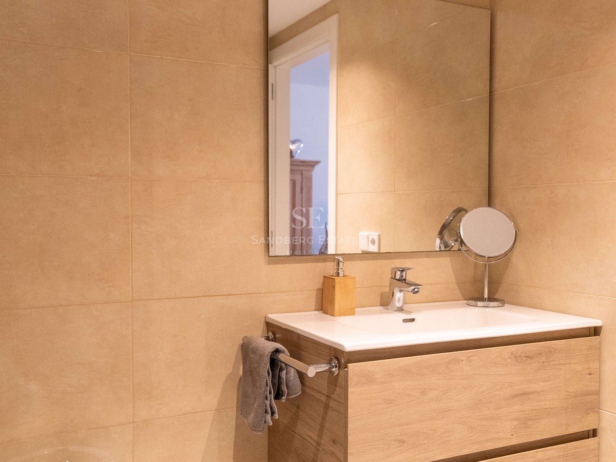 Salle de bain moderne avec meuble vasque suspendu en chêne, lavabo en céramique, grand miroir et carrelage beige.