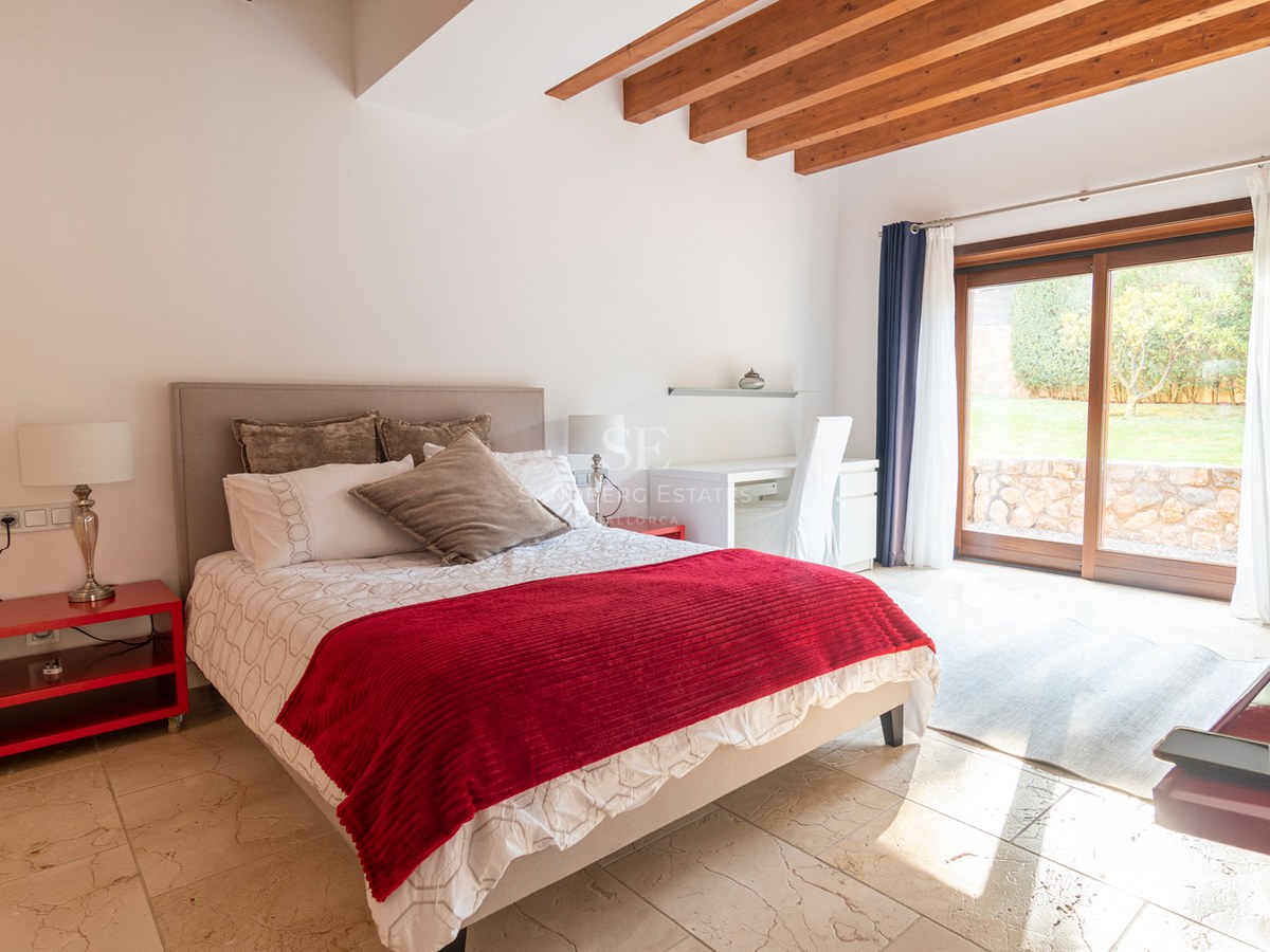 Chambre lumineuse avec accents rouges, sol en pierre, poutres en bois et baies vitrées donnant sur le jardin.