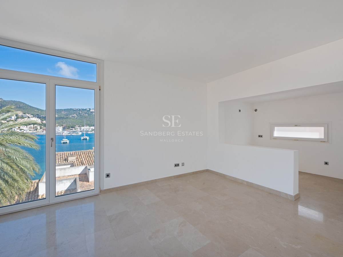 Empty minimalist room with cream marble floors and floor-to-ceiling windows overlooking a Mediterranean marina and mountains.