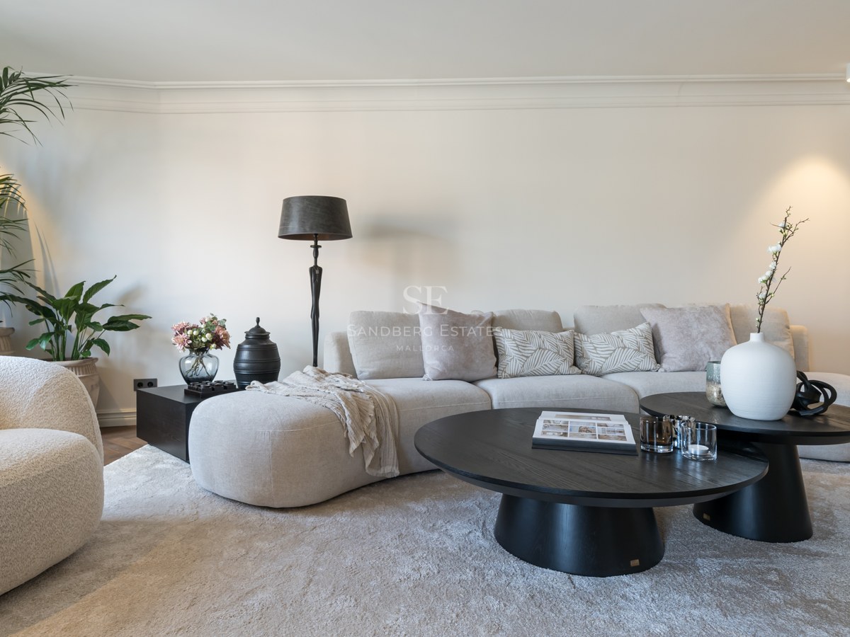 Salón moderno con sofá curvo beige, mesas de centro negras y alfombra mullida en una estancia luminosa y neutra.