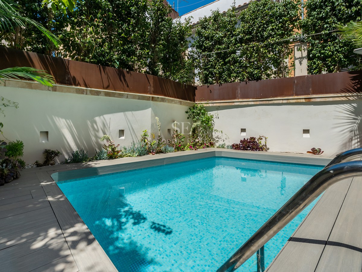 Small turquoise swimming pool with grey wooden decking surrounded by white walls and lush green plants.