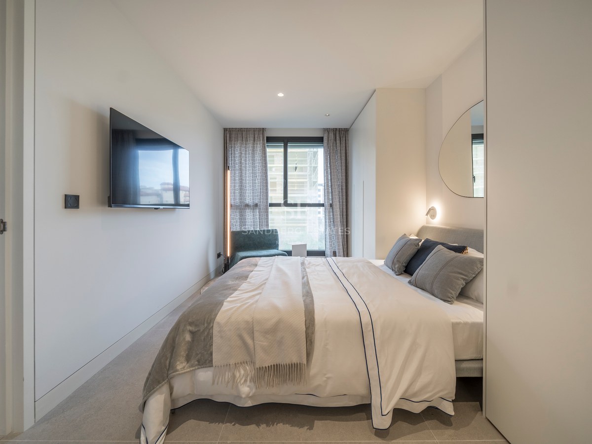 Chambre lumineuse avec TV murale, lit tapissé gris, miroir organique et grande fenêtre.