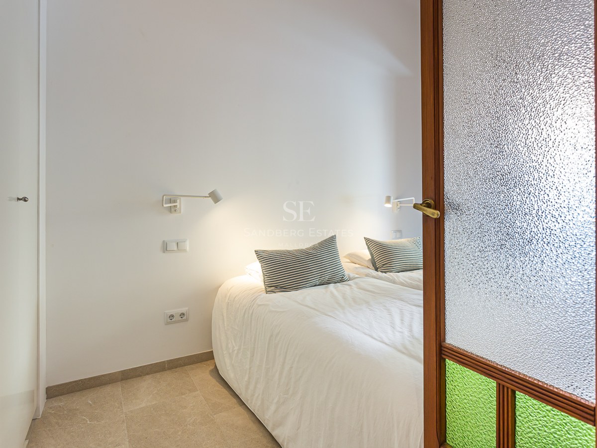 Dormitorio moderno visto a través de una puerta de madera con cristal texturizado, con cama blanca y lámparas de lectura.