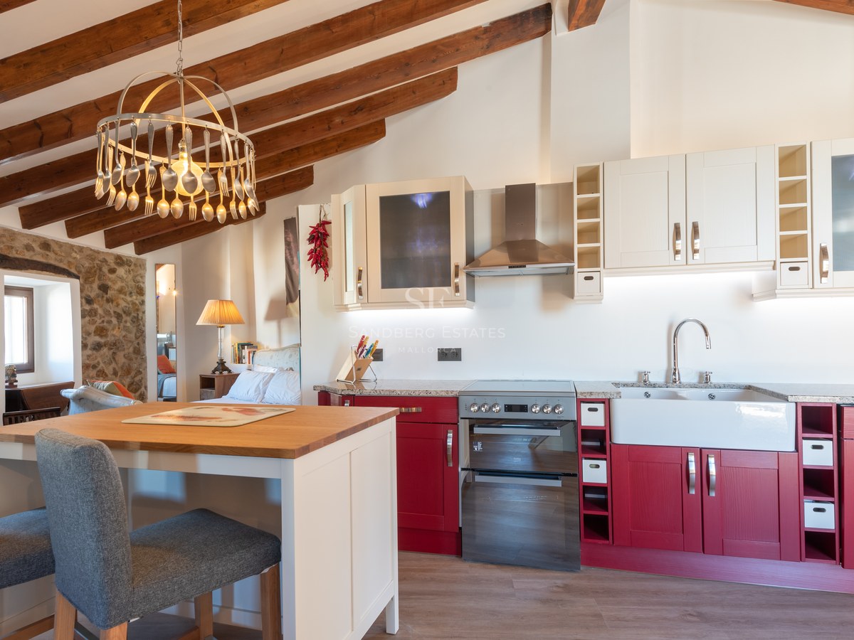 Cuisine avec placards rouges, poutres en bois apparentes et mur en pierre naturelle avec îlot central.