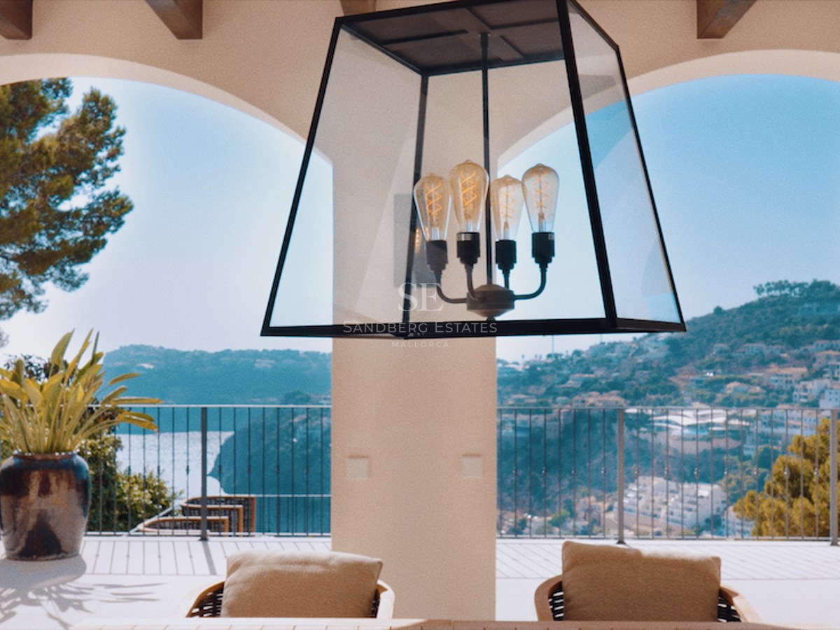View through white arches from a villa terrace overlooking the sea and green hills under a bright blue sky.