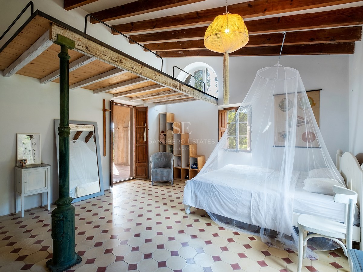 Grande chambre avec lit blanc, moustiquaire, mezzanine en bois, poutres apparentes et sol en carreaux décoratifs.