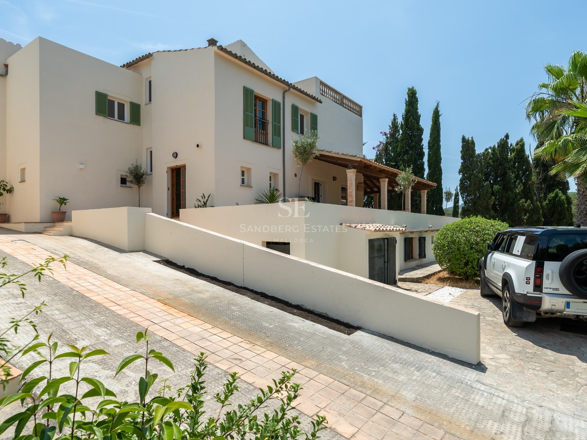Exterior view of a white multi-level Mediterranean villa with green shutters, a paved driveway, and palm trees.