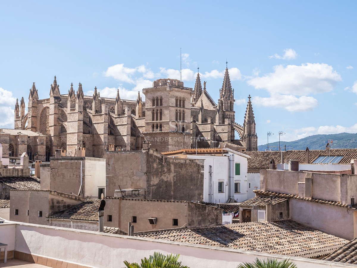 A large Gothic cathedral towers over the terracotta rooftops of a Mediterranean city under a blue sky.