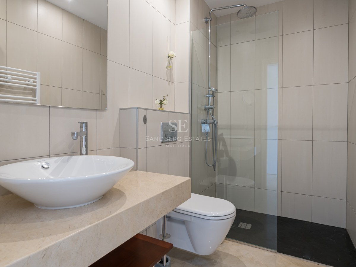 Baño moderno con azulejos beige, encimera de mármol, lavabo sobre encimera y ducha con mampara de cristal.