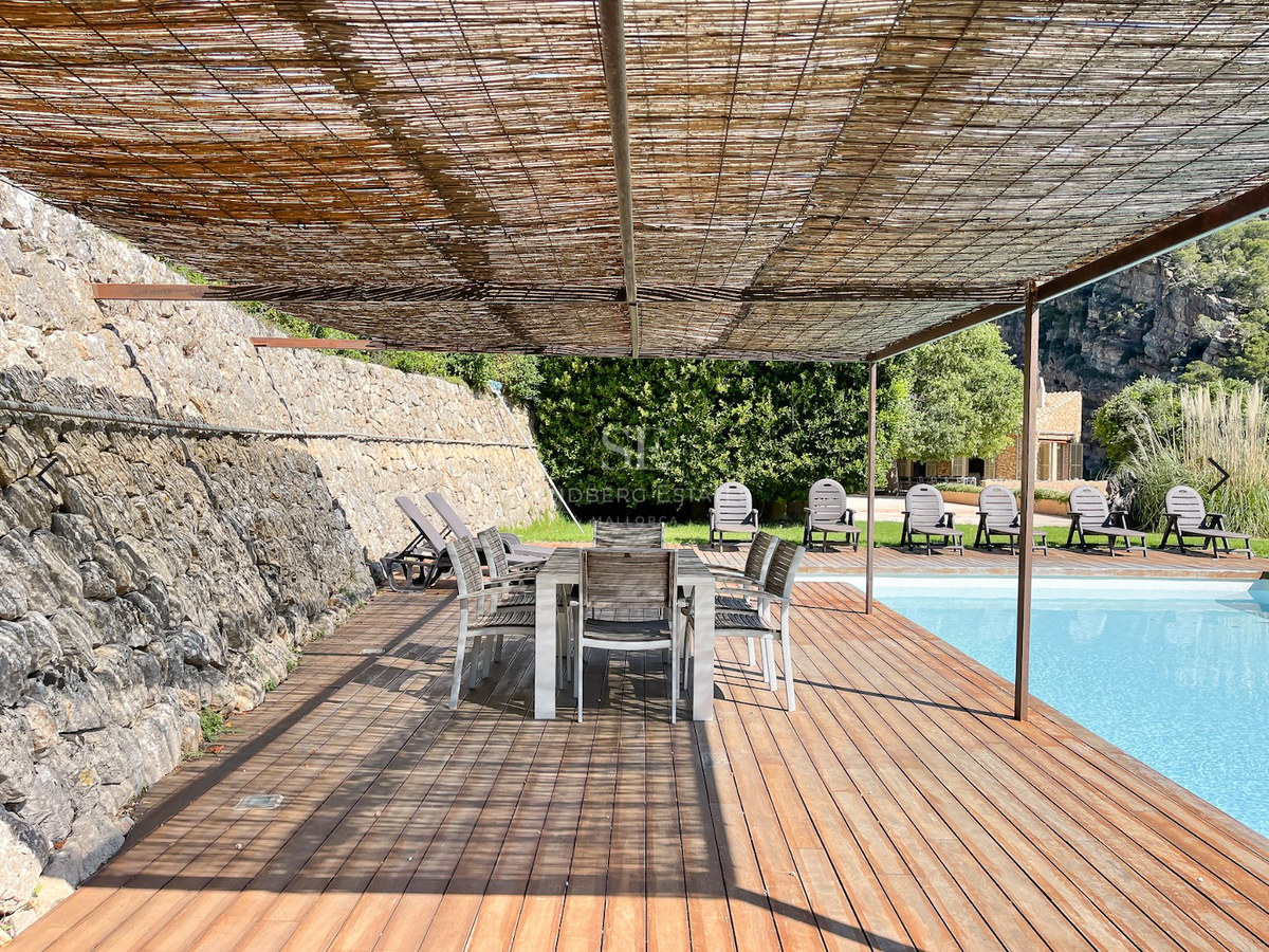 Terraza de madera junto a la piscina con comedor bajo pérgola de cañizo y muro de piedra natural.